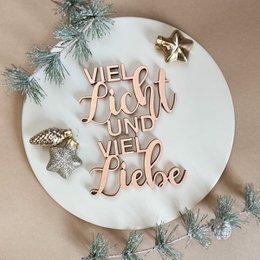 A round white decorative plate with the German text "Viel Licht und viel Liebe" (Lots of light and lots of love) written in rose gold lettering. The plate is decorated with pine branches and gold star-shaped ornaments.