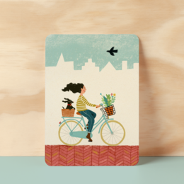 A postcard featuring a woman riding a bicycle through a town with buildings and a bird in the sky.
