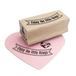 A wooden rubber stamp with the text "Enjoy the little things" printed on it. The stamp is shown on a pink heart-shaped background.