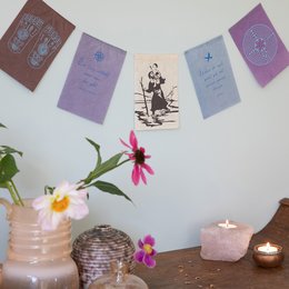 Colorful wishing flags with spiritual imagery and text, including "Life Journey" and "Wunschfähnchen | Lebensreise". The flags are displayed on a wall with flowers and candles, creating a serene and contemplative atmosphere.