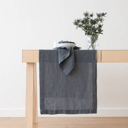 A long, rectangular linen table runner in a blue stone-washed color. The runner is shown draped over a wooden table, with a vase of greenery placed on top. The runner is available in multiple sizes, including 50 x 170 cm, 46 x 150 cm, 46 x 200 cm, and 46 x 250 cm.