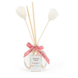 A clear glass reed diffuser bottle with a pink ribbon and floral accents. The bottle contains a liquid fragrance and is accompanied by several white reed sticks.