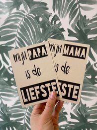 A greeting card with the text "Mijn papa is de liefste" (My dad is the sweetest) written on a handwritten sign against a tropical leaf background.