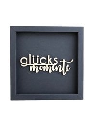 A wooden frame card with the text "MOMENTS OF HAPPINESS" in various colors including altrosa, taubengrau, taupe, eosin, aubergine, petrol, rehbraun, dunkelgrau, and schwarz.