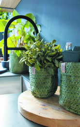 Olive-colored knitted paper bag with waterproof biodegradable liner, available in various sizes. Potted plants with green leaves and flowers on a wooden surface.