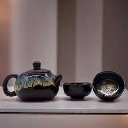 A black ceramic teapot set with a teapot and two cups, featuring a unique glaze pattern. The set is suitable for serving tea and has a traditional Asian-inspired design.
