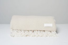 Herringbone-patterned throw blanket in a neutral cream color, featuring a soft, cozy texture and fringed edges.