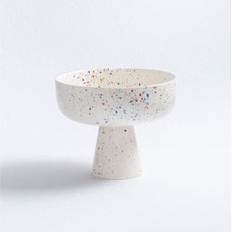 A round, white ceramic bowl with a pedestal base, featuring a speckled pattern of colorful dots throughout.