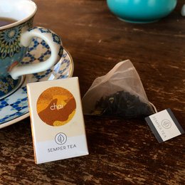 Organic black tea with spices in a pyramid tea bag. The tea bag features a circular design with a sun-like symbol. The tea is served in a ceramic cup with a saucer, alongside a turquoise ceramic bowl.