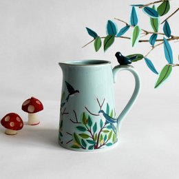 A ceramic jug with a floral and bird design, featuring a mint green color and a handle shaped like a bird.