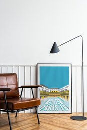 Framed wall art featuring a scenic landscape with a winding road, green hills, and a blue sky. The artwork has a minimalist, modern style with clean lines and a vibrant color palette.