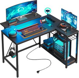 A large, L-shaped black carbon gaming desk with LED lighting. The desk features ample space for a computer setup, including a monitor, keyboard, and other gaming peripherals.