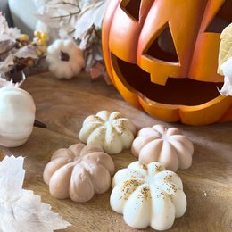 Orange pumpkin-shaped candle with assorted scented candle variants in pumpkin, vanilla, fig, maple, and witch's spell scents. Includes white pumpkin-shaped decorative elements.