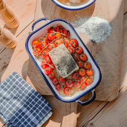 Rectangular enamel baking dish with a plain white interior and blue trim. The dish contains roasted tomatoes and a piece of fish or meat.