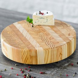 A round, wooden chopping block or cheese board made of larch wood, measuring approximately 25.4 cm in diameter and 5 cm in height. The surface has a natural, textured wood grain pattern and is topped with a small white cheese wedge garnished with fresh herbs and peppercorns.