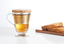 A clear glass tea set with 3 pieces, including a mug with a bee design, a stainless steel infuser, and a wooden coaster.