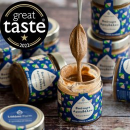 Creamy almond butter in a glass jar with a blue and white label. The jar is surrounded by other jars of the same product. The image also shows a Great Taste 2022 award logo and a vegan logo.