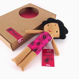 A plush Indian-inspired doll with a black hat, pink patterned dress, and wooden body. The doll is labeled "Rajani" and appears to be part of a baby doll collection.