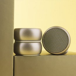 A round, metallic speaker in blue and gold colors. The speaker has a perforated mesh surface and a cylindrical shape, suitable for use as a phone holder.