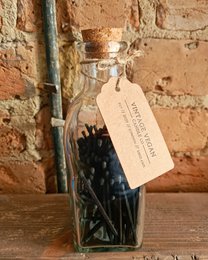 Black candle matches in a glass bottle. Matches are cylindrical in shape and black in color. The bottle has a cork stopper and a label attached.