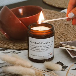 A round, tall candle in a brown glass container labeled "Grandma's Pumpkin Pie 155 g. Candle". The candle is scented and has a warm, cozy autumn-inspired aroma.