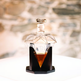 A tall, diamond-shaped glass decanter with a wooden base. The decanter contains a dark amber-colored liquid, likely liquor or wine. The decanter has a unique geometric design and appears to be a high-quality, decorative piece.