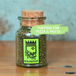 Herbal Mixture Mamma Mia! is a seasoning product in a glass jar with a cork lid. The jar contains a mixture of dried herbs and spices, likely used as a topping or seasoning for various dishes. The product label features a green logo with a crown and leaf design, indicating it is a natural or organic product.