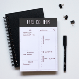 A black and white spiral-bound daily planner with the text "LET'S DO THIS!" printed on the cover. The planner contains a schedule layout with sections for tasks, appointments, and notes.