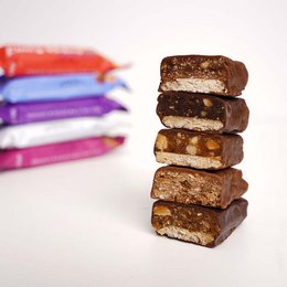 Lini's Bites bars are a variety of snack bars featuring flavors like almond cookie dough, salted peanut caramel, hazelnut nougat chocolate, cashew chocolate crunch, and coconut milk chocolate. The bars have a layered, rectangular shape and are wrapped in colorful packaging.