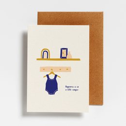 A postcard featuring a navy blue baby romper on a shelf with a rainbow and framed artwork. The text on the postcard reads "Happiness is in a little romper".