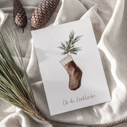 A6 greeting card with a watercolor illustration of a Christmas stocking on textured paper. The card features a pine branch and pine cones, creating a cozy, festive atmosphere.