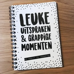 A black and white spiral-bound notebook with the text "I love unspoken & grappige momenten" (I love unspoken and funny moments) printed on the cover. The inside pages feature handwritten text in Dutch.