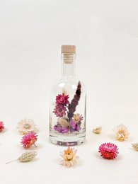 A glass bottle filled with dried fuchsia flowers, surrounded by other dried flowers in various colors.