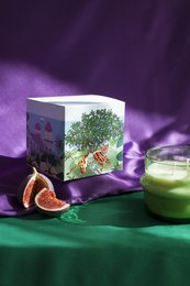 Tall, round, white scented candle with a fig and olive tree design on the packaging. The candle is displayed on a green cloth with a glass of green juice and a fresh fig.