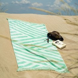 Large striped beach towel in mint green and white color, with a book and sunglasses on the sandy beach.