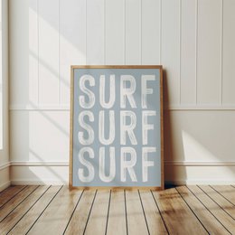 A framed poster with the text "SURF SURF SURF" in a minimalist, typographic design. The poster has a light blue background and is displayed against a white wall with wooden flooring.