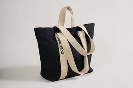 A black tote bag with a large white logo design, made of durable canvas material. Suitable for everyday use as a shoulder bag or shopping bag.