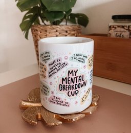 A white ceramic mug with the text "My Mental Breakdown Mug" printed on it, along with various mental health-related phrases. The mug is placed on a wooden surface with a potted plant in the background.