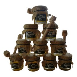 A rustic honey jar with a wooden spoon and burlap accents, surrounded by other honey jars in a stacked arrangement. The jars feature various honey flavors and varieties.