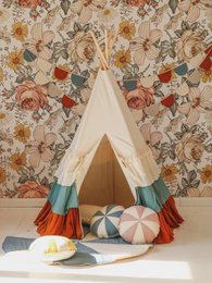 A colorful teepee-style play tent with a patterned fabric exterior, featuring frills and a brown-beige mat. The tent is accompanied by decorative pillows and balls, creating a whimsical and inviting play space for children.