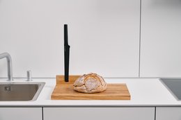 Wooden bread board with a loaf of bread on a kitchen counter. The bread board is rectangular in shape and made of light-colored wood. A black knife is placed next to the bread.