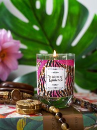 A tall, round scented candle with a tiger-striped pattern and the text "The Jungle Collection" printed on the label. The candle is surrounded by tropical leaves and flowers, creating a lush, natural setting.