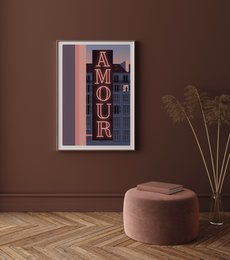 Framed wall art with the text "AMOR" in a retro-style design, featuring a pink and purple color scheme. The artwork is displayed in a minimalist living room setting with a pink pouf and dried plants.