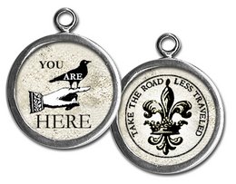 A round pendant with a black silhouette of a bird and the text "YOU ARE HERE" on a beige background. The reverse side features a fleur-de-lis symbol and the text "MAKE THE ROAD LESS TRAVELED".