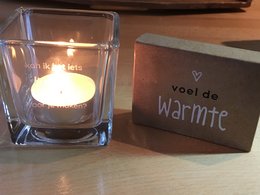 Glass tea light holder with the text "Kan ik het iets LICHTER voor je maken?" and "De liefde blijft..." printed on it. The holder contains a lit tea light candle.