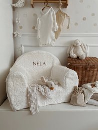 Soft, plush white chair with embroidered name "Nela" for a child's room. Includes matching accessories like a stuffed animal and basket.