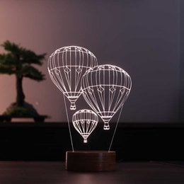 A decorative 3D LED lamp featuring two hot air balloons against a dark background with a bonsai tree silhouette. The lamp has a wooden base and emits a warm, ambient light.