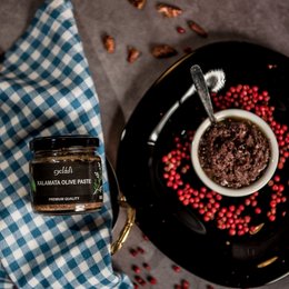 Olive paste in a glass jar, with kalamata olives, chili peppers, and capers as product variants. The paste is dark in color and appears to have a thick, spreadable texture. The image also shows a checkered cloth, bread, and other ingredients, suggesting this olive paste may be used as a spread or condiment.