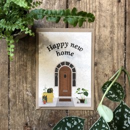 A postcard with the text "Happy new home!" and an illustration of a wooden door with plants around it, set against a wooden background with greenery.