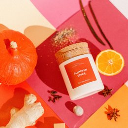A pumpkin spice scented candle in a glass jar with a cork lid. The candle is surrounded by autumn-themed decorations like a pumpkin, orange slices, and spices.
